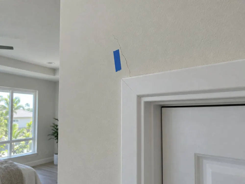 A hairline drywall crack above a door frame in a newer home, marked with blue tape.