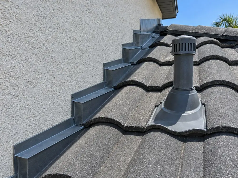 Concrete roof tiles meeting a stucco wall, with step flashing and a vent pipe.