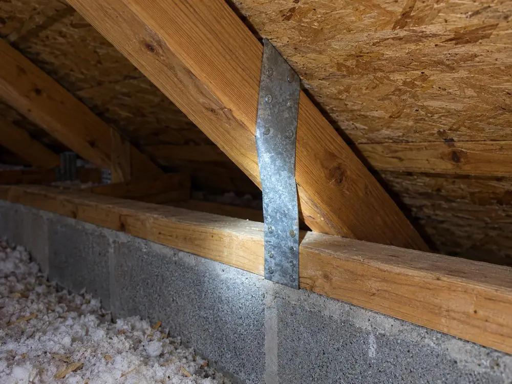 A metal roof-to-wall strap connecting a roof truss to a block wall in an attic.